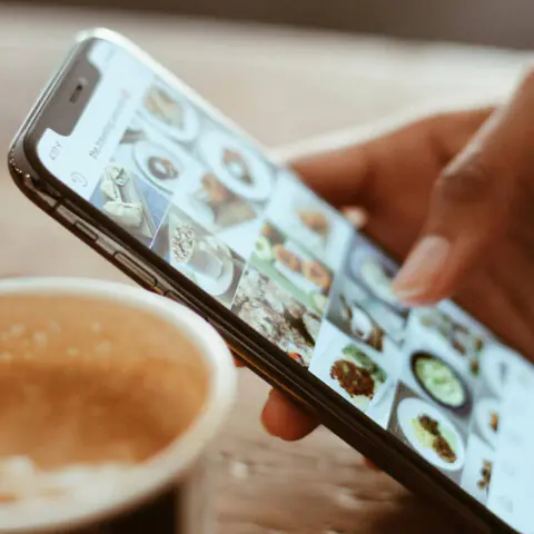 Light brown-skinned hands holding a mobile smartphone showing an Instagram grid of food-related images. Photo by Kerde Severin on Pexels.