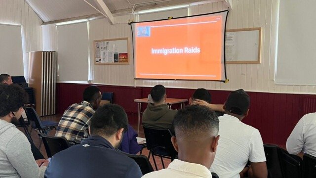 Refugees are seated in front of a projector screen watching a presentation about immigration raids.