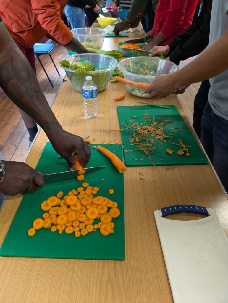 A man with arm tattoos chops brightly coloured orange carrots at the front of a line of other people chopping salad and vegetables together at long table.