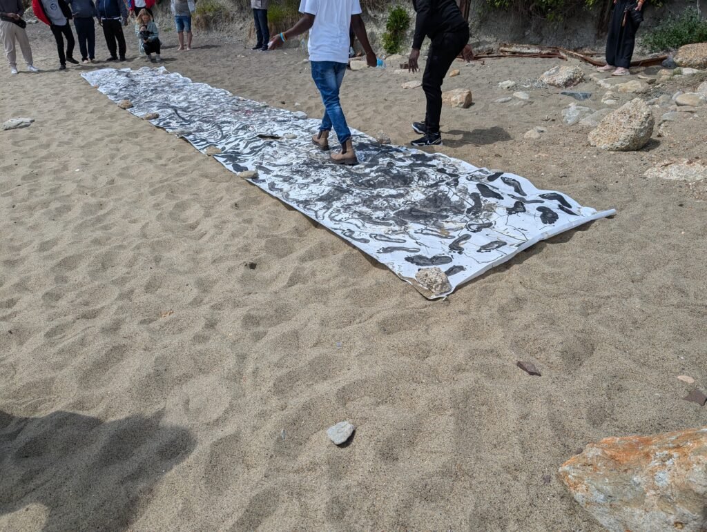 A man walks along a large scroll of paper decorated with black footprints on a sandy beach towards a woman kneeling down with a camera filming him. A crowd of people stand watching.