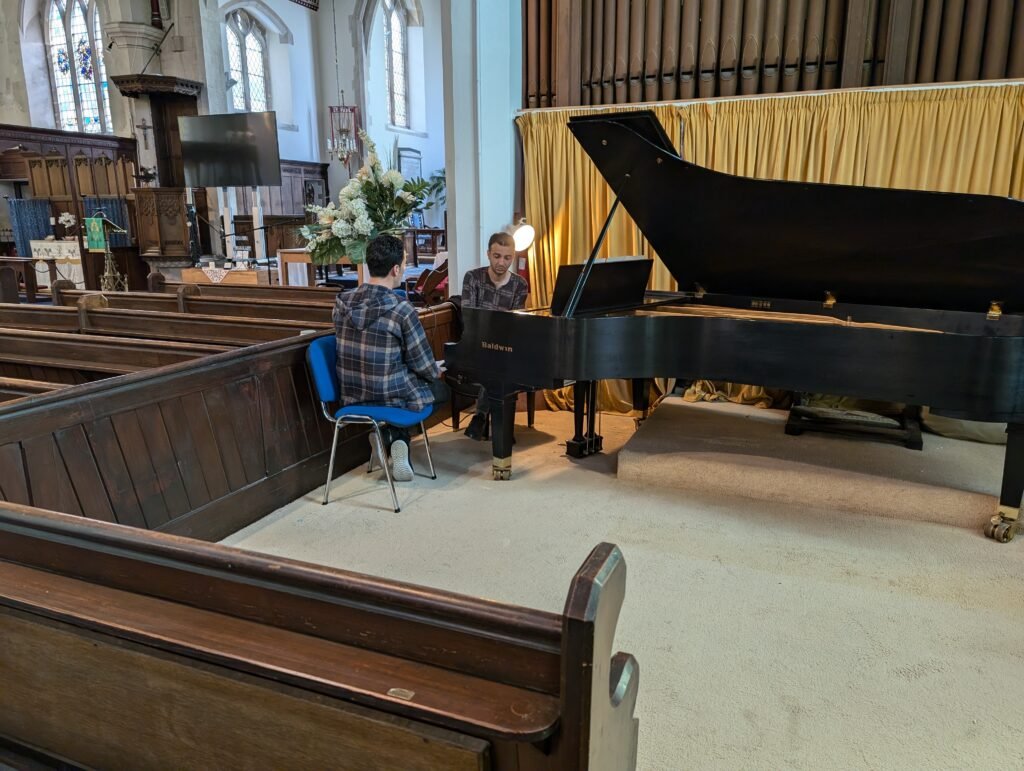 An Iranian man is seated behind a grand piano playing while another man faces him singing. They are seated in front of an organ in a traditional English church.