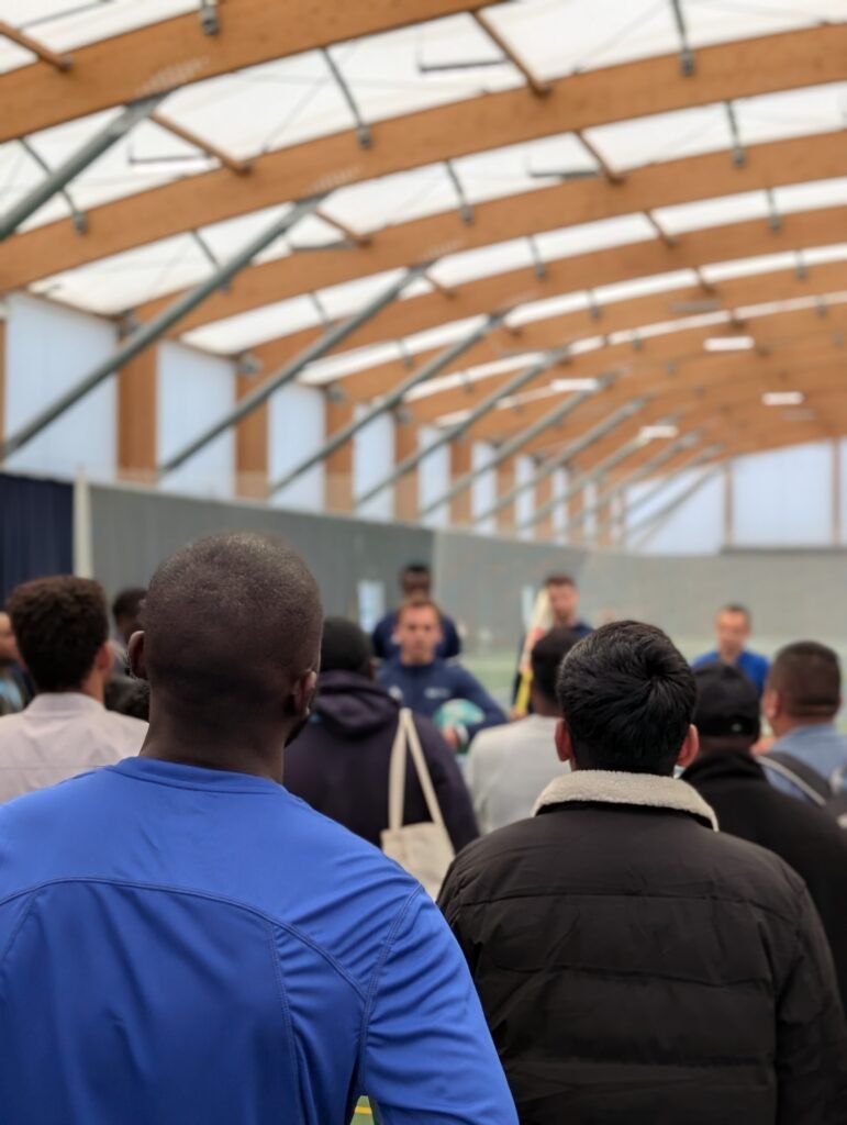 A man in a blue sports top and a man in a black jacket face away from the camera, amongst a large group of men of different ethnicities, listening to coaches in a vaulted sports hall giving instructions.