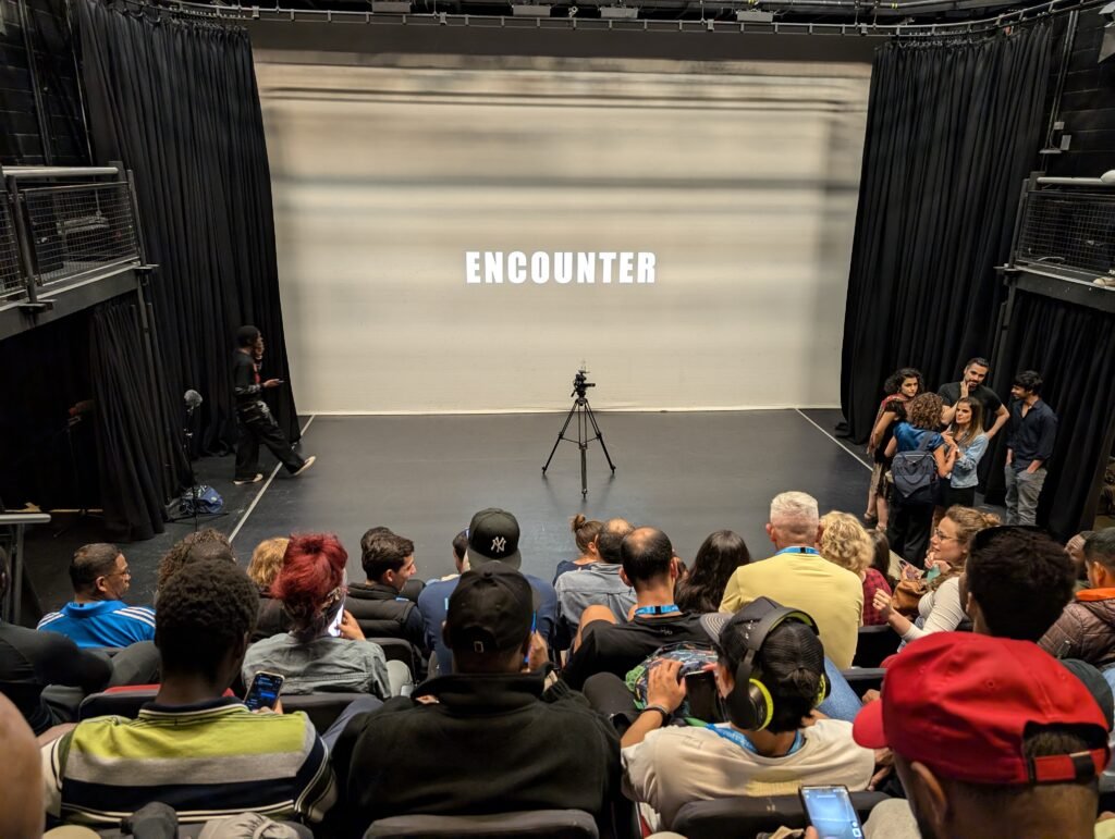 In a theatre, an audience of men and women await the screening of ENCOUNTER. This word is projected in caps onto the screen. There is a camera pointed towards the audience in the middle of the stage filming them.