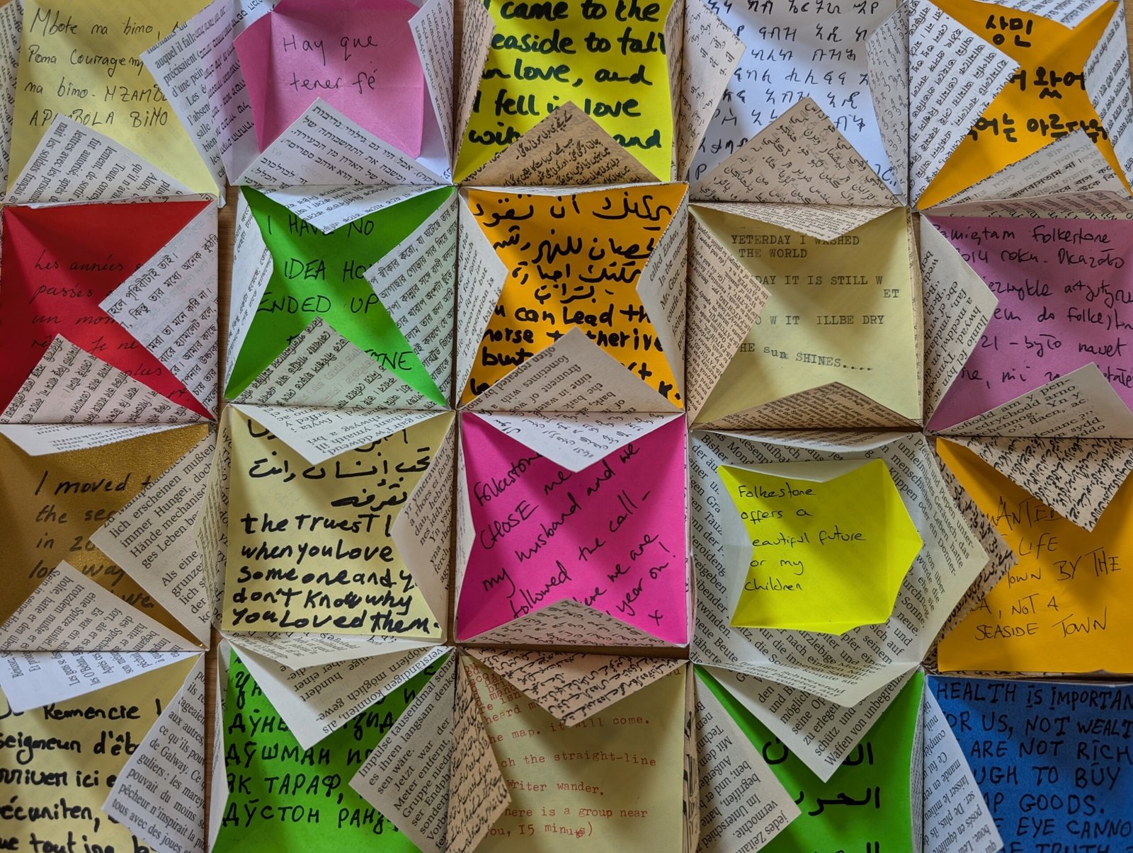 A set of 20 brightly coloured post-it notes with handwritten messages in different languages are arranged in a grid. Each post it note is stuck inside the page of a book folded around the post it note like a small envelope.